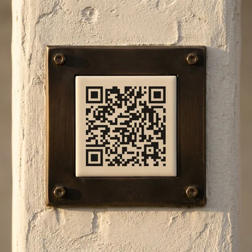 A brass plate holding a printed QR code, bolted to a plastered gate pillar in evening light.